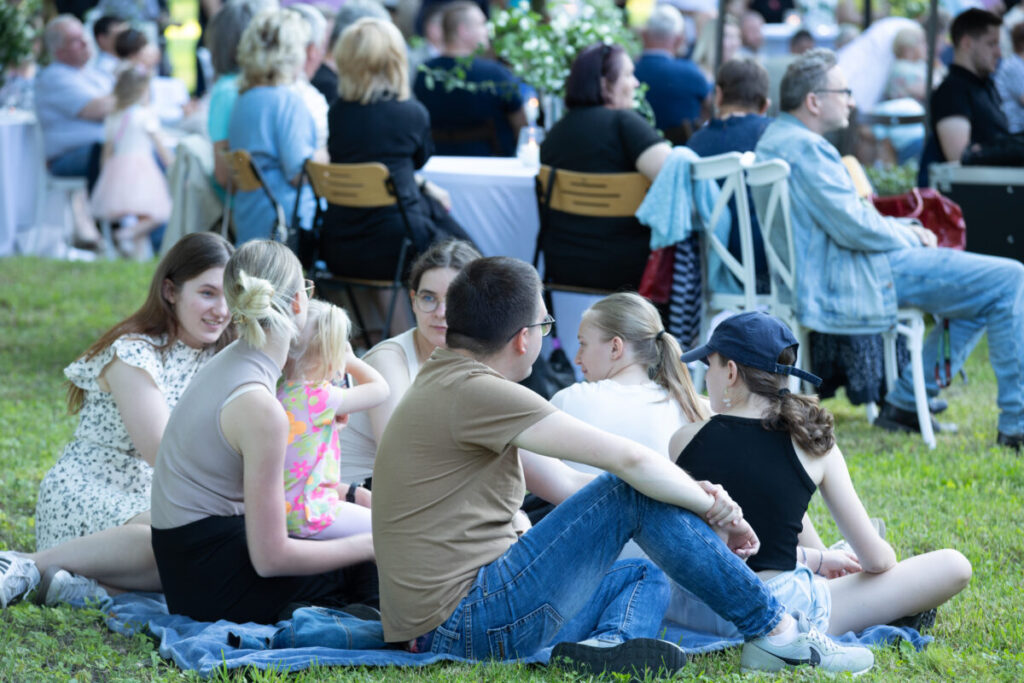 Grupa osób siedzi na trawie na kocu podczas pikniku. Na pierwszym planie młodzi ludzie, w tle widoczne stoły i krzesła. Jasne kolory ubrań i zielona trawa dominują na zdjęciu.