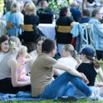 Grupa osób siedzi na trawie na kocu podczas pikniku. Na pierwszym planie młodzi ludzie, w tle widoczne stoły i krzesła. Jasne kolory ubrań i zielona trawa dominują na zdjęciu.
