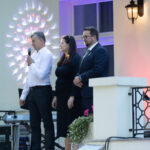 Trzy osoby na jasnym balkonie, jeden mężczyzna mówi do mikrofonu. Tło zdobią kolorowe światła. Osoby w eleganckich strojach.