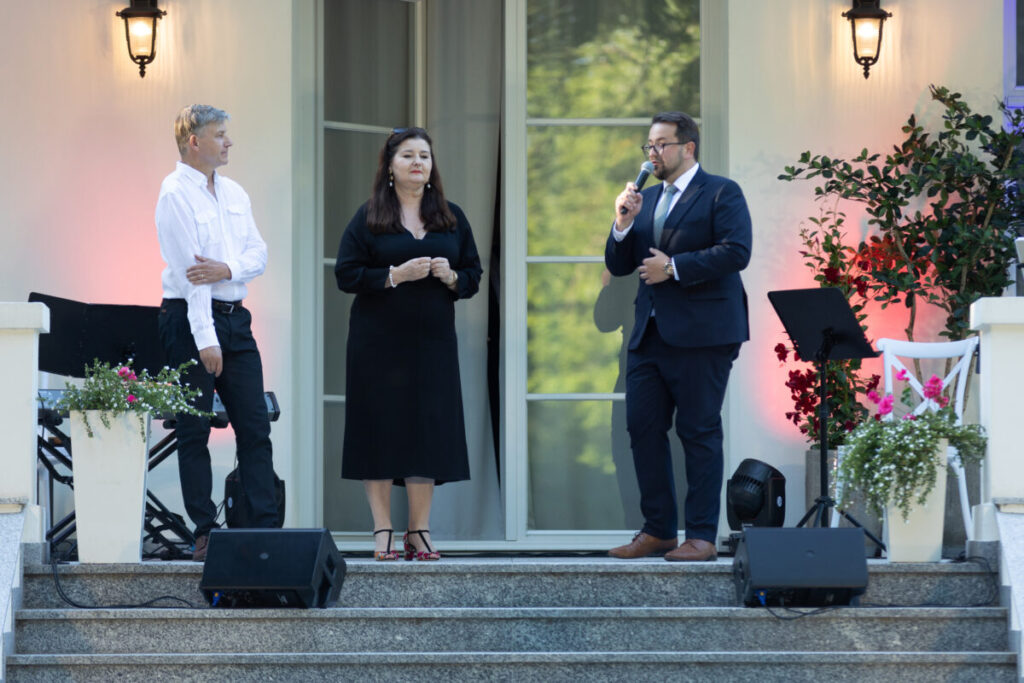 Na zdjęciu trzy osoby na schodach przed eleganckim budynkiem. Kobieta w czarnej sukni i dwóch mężczyzn. W tle jasna elewacja i rośliny.