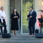 Na zdjęciu trzy osoby na schodach przed eleganckim budynkiem. Kobieta w czarnej sukni i dwóch mężczyzn. W tle jasna elewacja i rośliny.