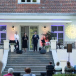 Trzy osoby stoją na schodach przed białym budynkiem z czerwonym dachem. W tle kolorowe światła i rośliny. Publiczność siedzi przodem.