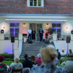 Na zdjęciu scena przed budynkiem z czerwoną dachówką. Troje ludzi przemawia przed widownią. W tle efekty świetlne i rośliny.