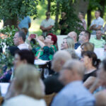 Grupa ludzi siedząca na zewnątrz przy stołach. Część osób robi zdjęcia telefonem. Zielone drzewa w tle.