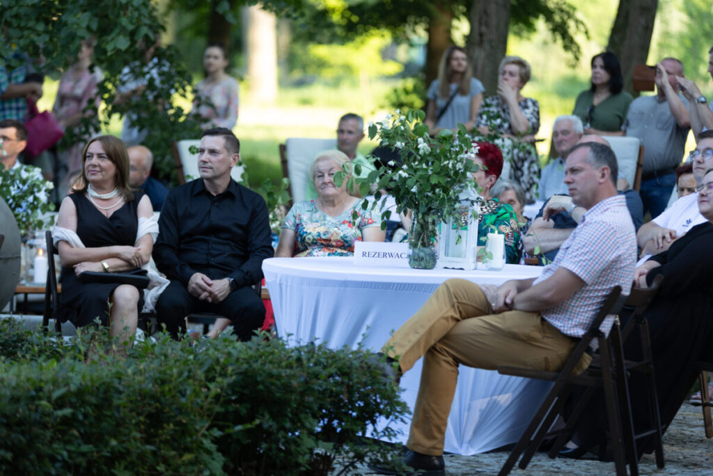 Grupa ludzi siedzi przy stole z białym obrusem na zewnątrz. Na stole są zielone rośliny i napis Rezerwacja. W tle zielone drzewa i więcej osób.