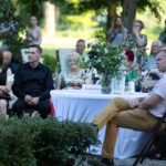 Grupa ludzi siedzi przy stole z białym obrusem na zewnątrz. Na stole są zielone rośliny i napis Rezerwacja. W tle zielone drzewa i więcej osób.