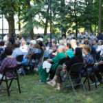 Ludzie siedzą na krzesłach w parku, otoczeni drzewami i zielenią. Ubrania mają różnorodne kolory, przeważają jasne odcienie. W tle widać inne osoby oraz namioty.