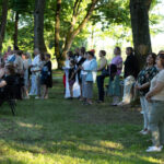 Grupa ludzi stoi na trawie, otoczona drzewami. Mają na sobie letnie ubrania w jasnych kolorach. W tle widać zielone liście.