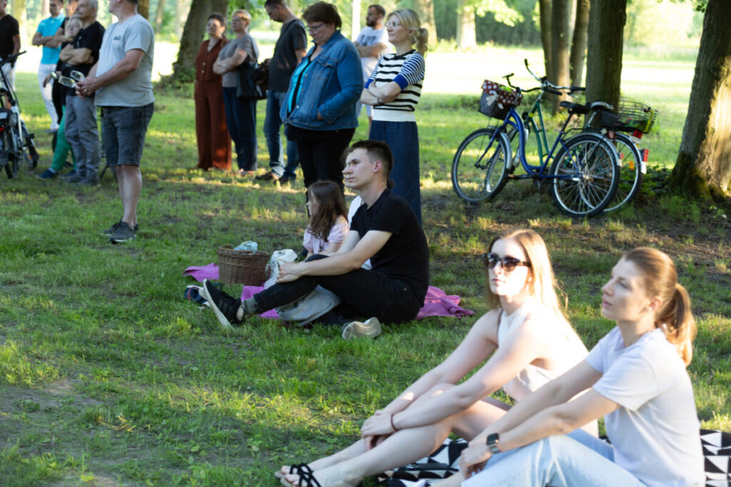 Grupa ludzi słucha na trawie w parku, w tle drzewa i rowery. Dominują kolory zieleni i odcienie ubrań. Słońce rzuca cienie.