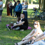 Grupa ludzi słucha na trawie w parku, w tle drzewa i rowery. Dominują kolory zieleni i odcienie ubrań. Słońce rzuca cienie.