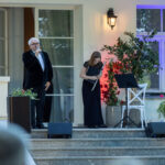 Na schodach występują mężczyzna z mikrofonem i kobieta z fletem. Jasne kolory elewacji, roślinność oraz oświetlenie. W tle okno i drzwi.