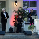 Mężczyzna w czarnym garniturze śpiewa na scenie z muzykami w tle. Tło w jasnych barwach z dekoracyjnym oświetleniem. Obok zielone rośliny w donicach.