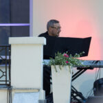 Mężczyzna przy klawiaturze na jasno oświetlonym tle. Obok biała donica z kwiatami. W tle okno z fioletowym światłem.