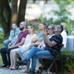 Ludzie siedzą na krzesłach na zewnątrz, w zielonym parku. Jeden mężczyzna robi zdjęcie telefonem. W tle drzewa i biały namiot.