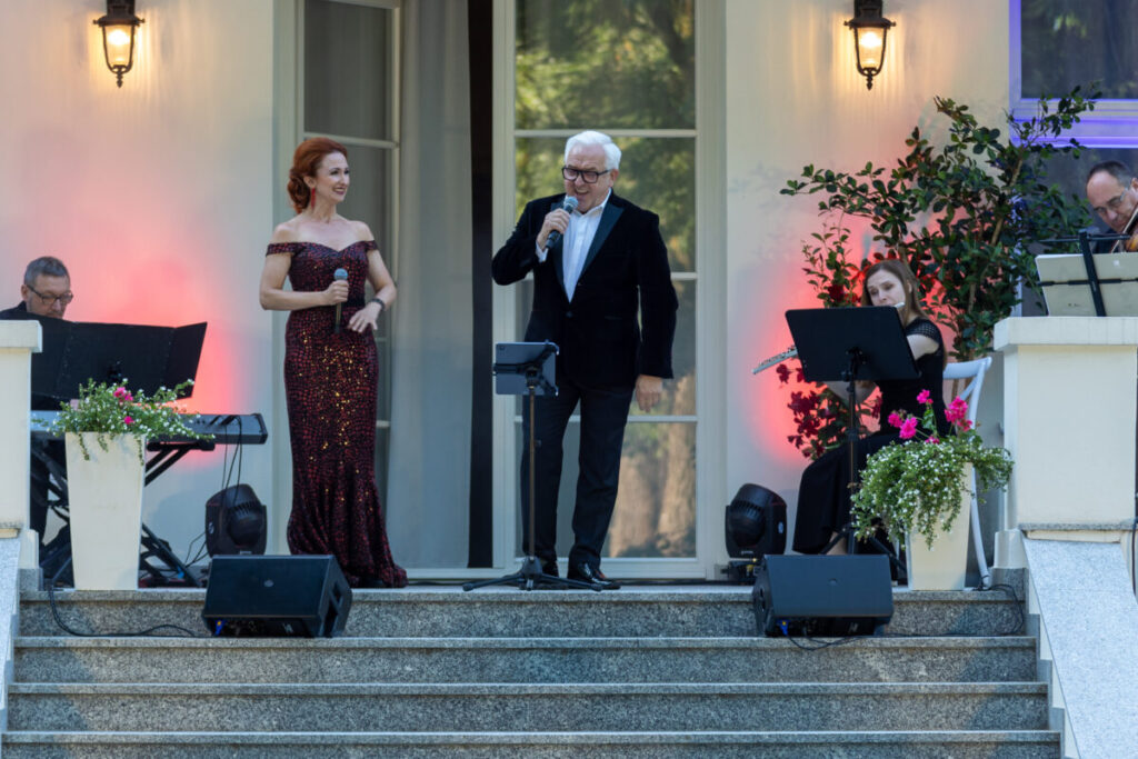 Na zdjęciu jest koncert na świeżym powietrzu. Kobieta w ciemnej, błyszczącej sukni i mężczyzna w czarnym garniturze śpiewają. Tło to jasna ściana i zieleń, z czerwonymi światłami.