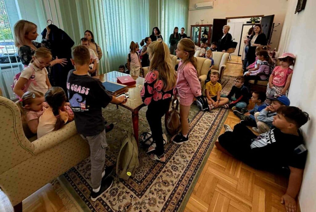 Grupa dzieci w różnorodnych kolorowych ubraniach siedzi i stoi w salonie z zielonymi zasłonami oraz drewnianą podłogą. Dzieci wokół stołu, na kanapach i dywanie, otoczone jasnymi ścianami. W tle dorośli obser