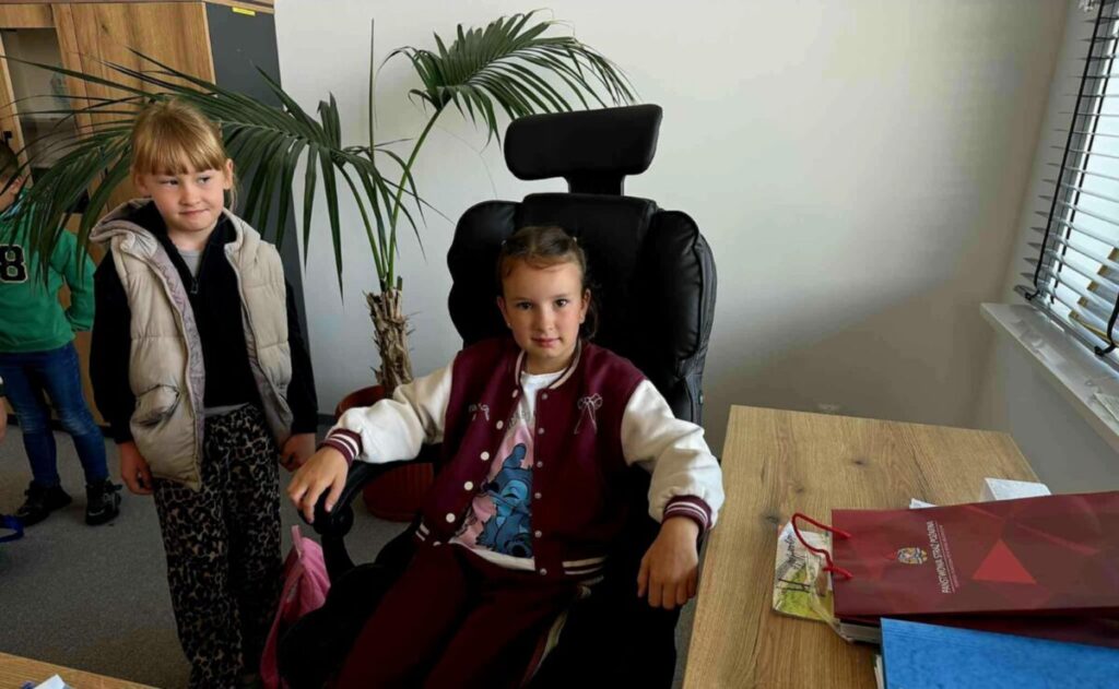 Dwie dziewczynki w biurze, jedna siedzi na czarnym fotelu. Obok stoi z palmą w tle. Drewniane biurko z teczką po prawej.