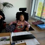 Dziewczynka w różowej bluzie siedzi na krześle biurowym. Na biurku znajduje się klawiatura i dokumenty. W tle okno z żaluzjami i roślina.