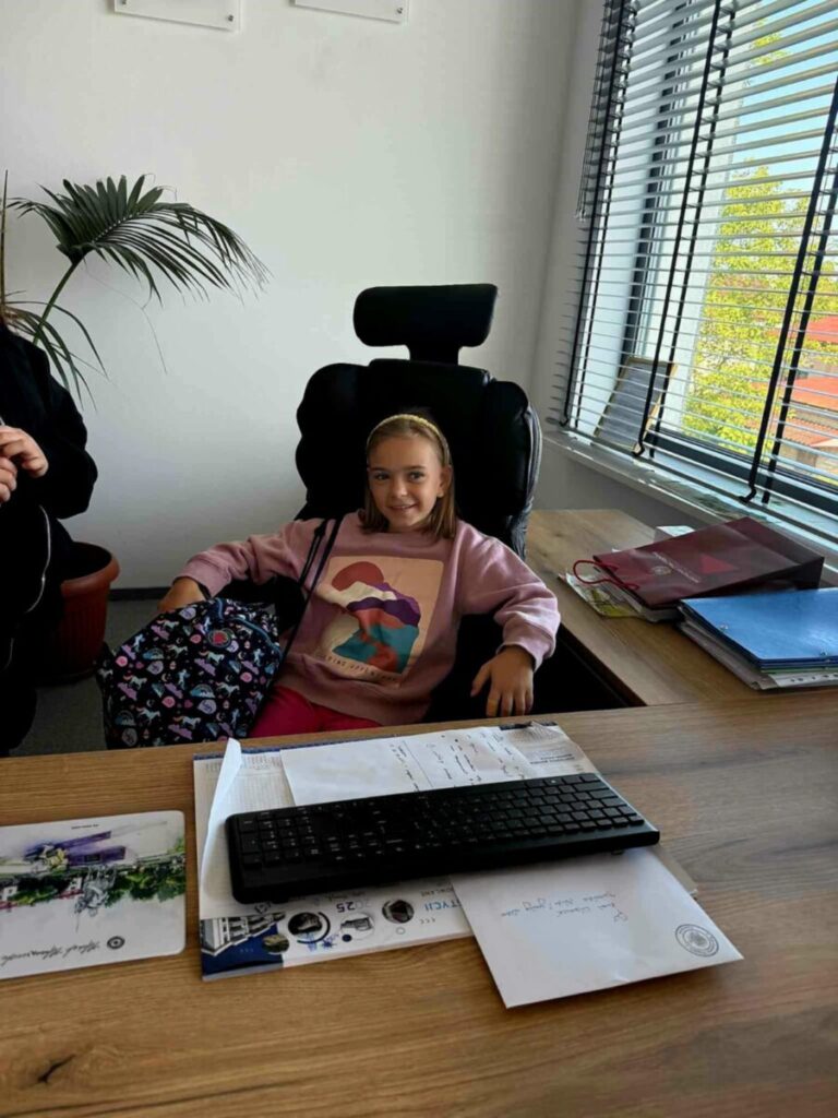 Dziewczynka w różowej bluzie siedzi na krześle biurowym. Na biurku znajduje się klawiatura i dokumenty. W tle okno z żaluzjami i roślina.