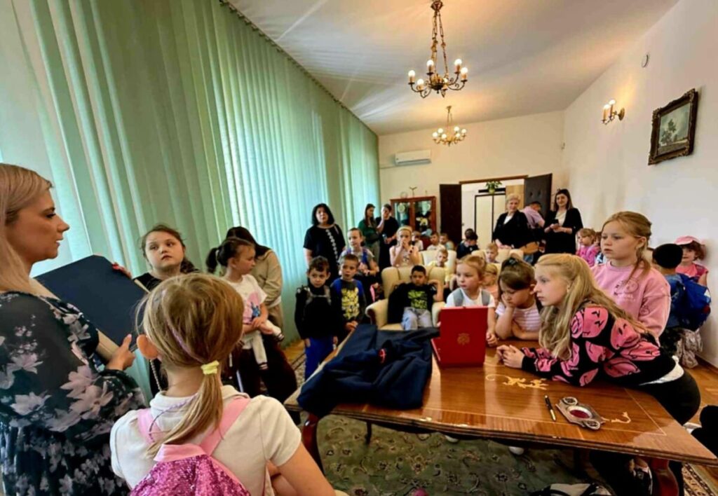 Grupa dzieci i dorosłych w pokoju z zielonymi zasłonami, drewnianym stołem i żyrandolem. Dzieci słuchają kobiety. Atmosfera edukacyjna.