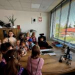 Grupa dzieci stoi wokół biurka w biurze z dużym oknem i roletami. W tle widoczne są rośliny i plakaty na ścianie. Na biurku leżą dokumenty i komputer.