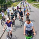 Grupa rowerzystów jedzie ulicą w słoneczny dzień. Widoczne zielone krzewy i domy w tle. Różnorodne stroje, kaski i plecaki.