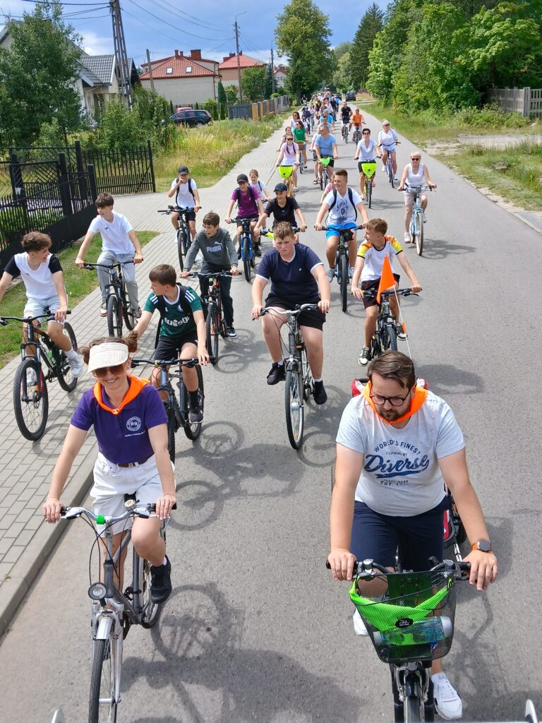 Grupa rowerzystów jedzie ulicą w słoneczny dzień. Widoczne zielone krzewy i domy w tle. Różnorodne stroje, kaski i plecaki.