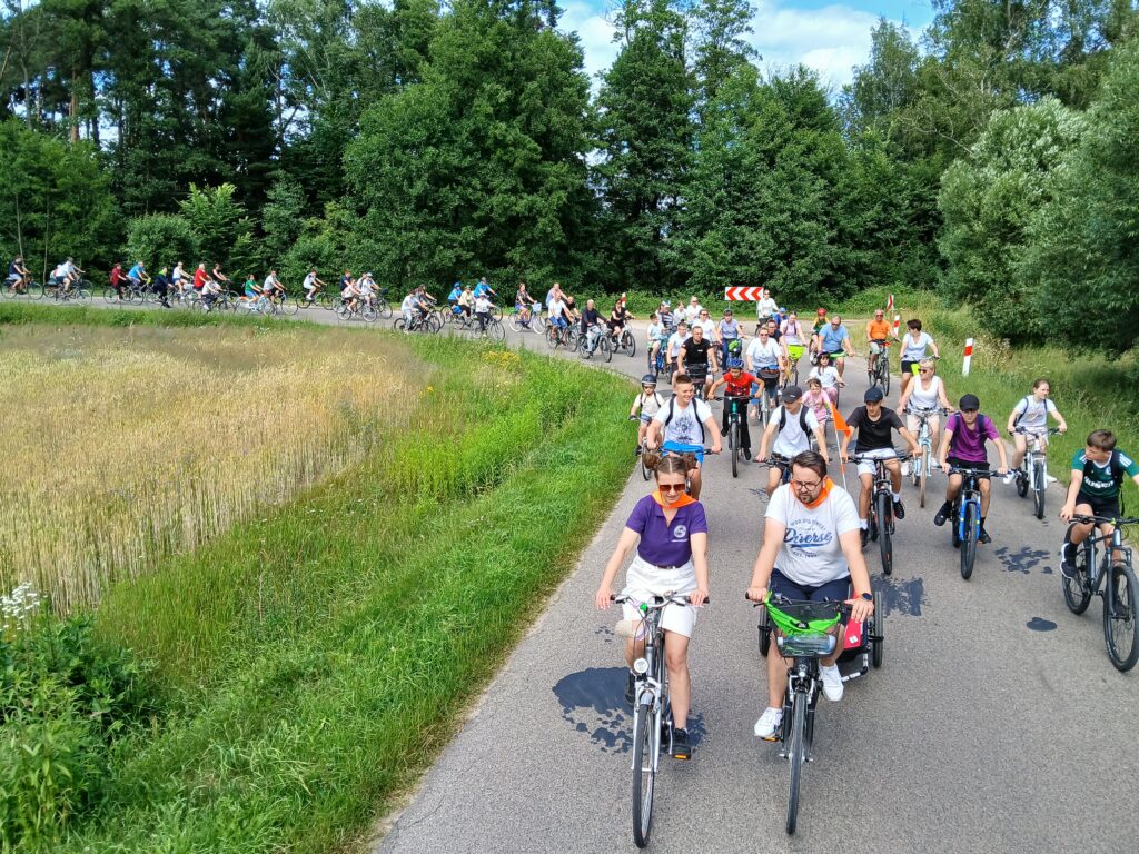 Grupa rowerzystów na wiejskiej drodze otoczonej zielenią, słońce świeci jasno, w tle las i niebo.