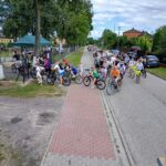 Grupa rowerzystów na ścieżce, otoczona zielenią i domami, pod niebieskim, zachmurzonym niebem. Alpinizm, czerwony bruk na drodze.