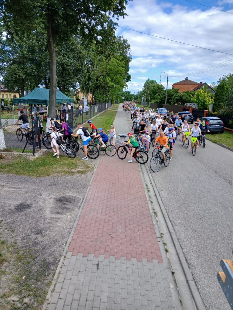 Grupa rowerzystów na ścieżce, otoczona zielenią i domami, pod niebieskim, zachmurzonym niebem. Alpinizm, czerwony bruk na drodze.