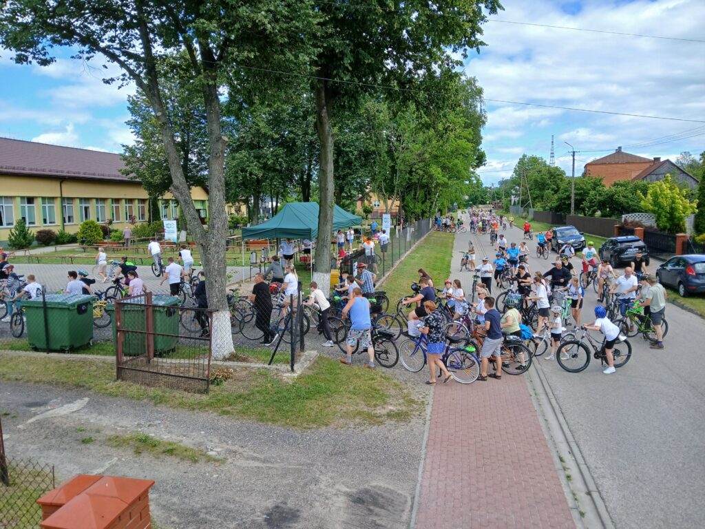 Grupa ludzi na rowerach stoi na ulicy. W tle budynek, otoczenie zielenią. Niebieskie niebo z białymi chmurami.