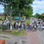 Grupa ludzi na rowerach stoi na ulicy. W tle budynek, otoczenie zielenią. Niebieskie niebo z białymi chmurami.