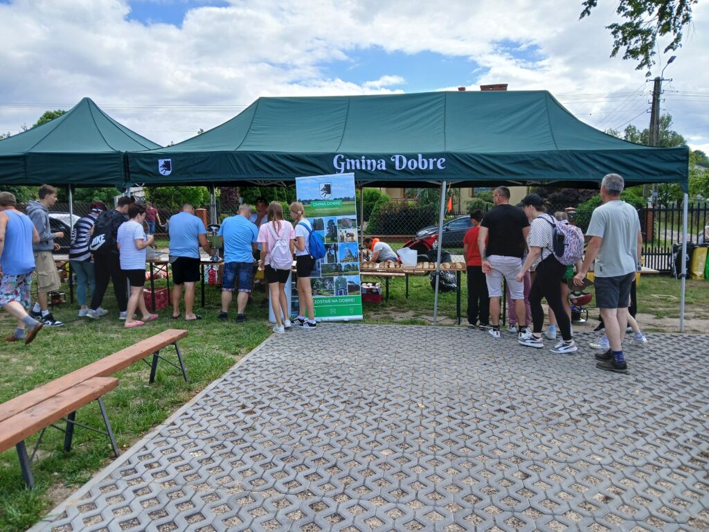 Ludzie stoją w kolejce pod zielonym namiotem na targu. W tle widoczne są chmury i trawa. Obok wisi baner Gmina Dobre.