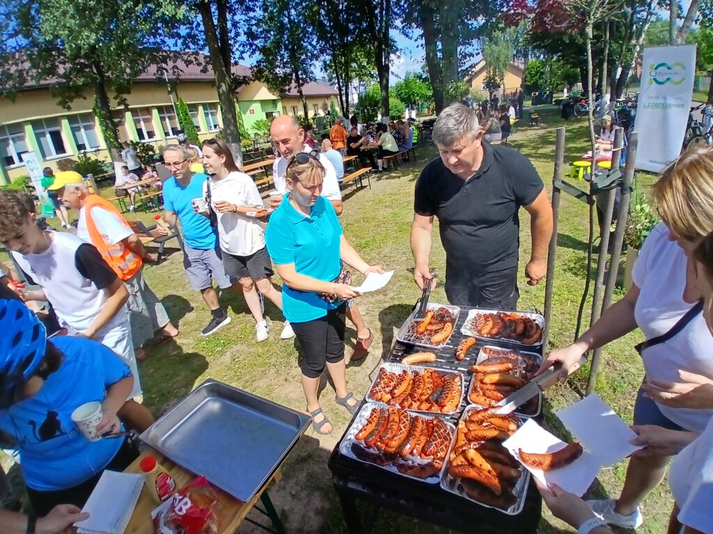 Grupa ludzi na pikniku próbuje kiełbasek z grilla. Tło stanowi zielony teren z drzewami i budynkami. Pogoda jest słoneczna, co podkreślają jasne kolory ubrań uczestników.