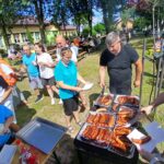 Grupa ludzi na pikniku próbuje kiełbasek z grilla. Tło stanowi zielony teren z drzewami i budynkami. Pogoda jest słoneczna, co podkreślają jasne kolory ubrań uczestników.