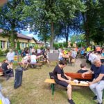 Na zdjęciu widać ludzi siedzących na ławkach pod drzewami na pikniku. Tło stanowi budynek z kremową elewacją. Dominuje zielona i niebieska kolorystyka.