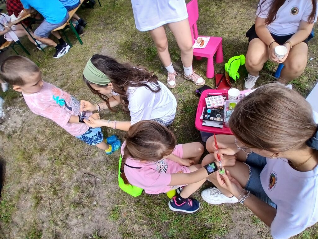 Grupa dzieci i dorosłych na trawie maluje farbami. Ubrania mają jasne kolory, a w tle widać stół z osobami. Atmosfera plenerowa.