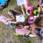 Grupa dzieci i dorosłych na trawie maluje farbami. Ubrania mają jasne kolory, a w tle widać stół z osobami. Atmosfera plenerowa.