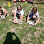 Trójka dzieci w jasnych koszulkach czyta gazety na trawie. Za nimi widać ludzi i kolorowe pachołki. Słoneczny dzień.