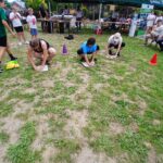 Na trawie klęczą trzy osoby, skupione na zadaniu, wokół ludzi. Na drugim planie znajduje się zielony namiot i stoły. Tło jest zielone i żywe, z czerwonym pachołkiem po prawej.