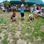 Grupa ludzi na trawie, biorących udział w aktywności. Zielony namiot w tle, kolorowe stożki na ziemi. Dzień słoneczny, atmosfera piknikowa.