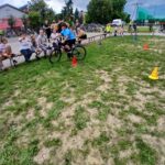 Na zdjęciu widać osobę na rowerze, manewrującą między kolorowymi pachołkami na trawie. W tle siedzą ludzie na ławkach, niebo jest pochmurne. Otoczenie jest zielone z kilkoma drzewami.