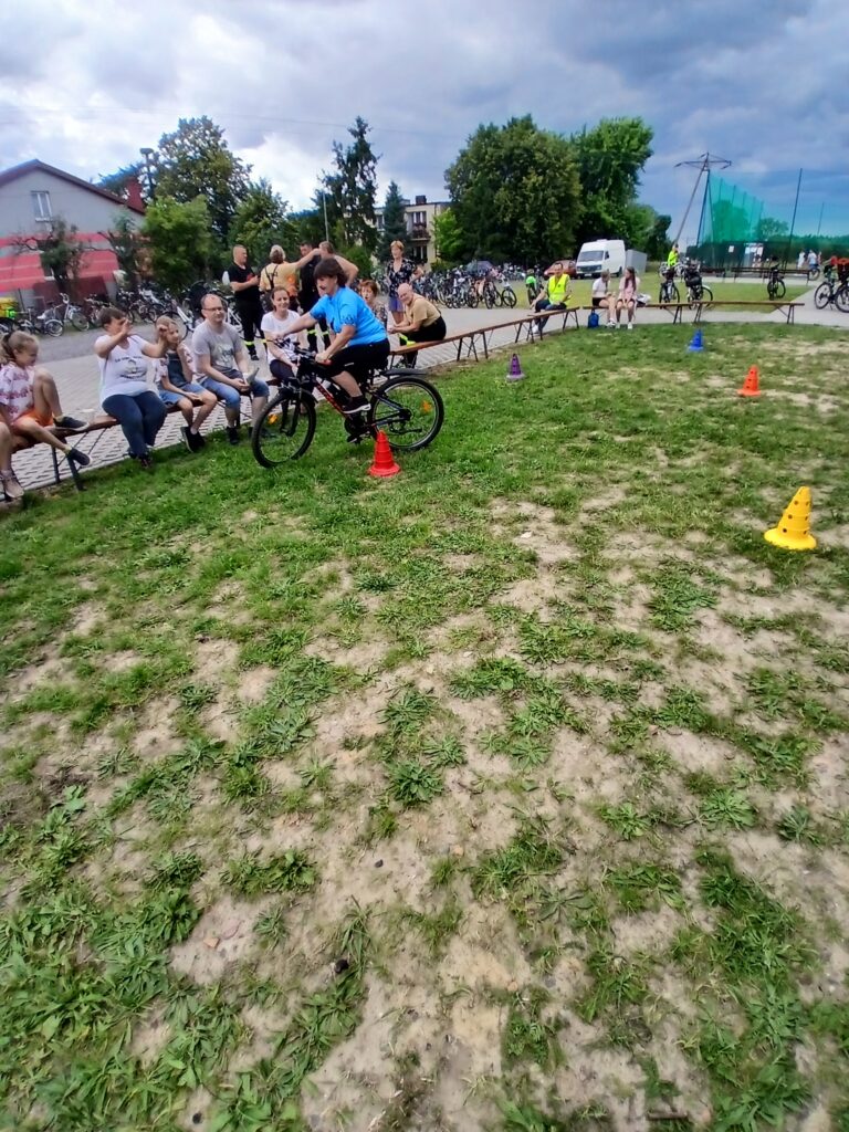 Na zdjęciu widać osobę na rowerze, manewrującą między kolorowymi pachołkami na trawie. W tle siedzą ludzie na ławkach, niebo jest pochmurne. Otoczenie jest zielone z kilkoma drzewami.