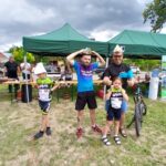 Cztery osoby pozują pod zielonymi namiotami na zewnątrz. Dwie z nich mają papierowe kapelusze, a dwoje dzieci nosi stroje rowerowe. Tło stanowią drzewa i stoiska.