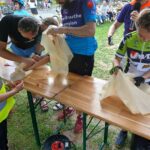 Na zdjęciu kilka osób szyje przy drewnianych stołach na świeżym powietrzu. Ubrani są kolorowo, wokół widać rowery. W tle grupka ludzi obserwuje wydarzenie.