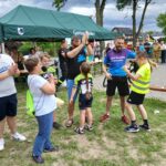 Grupa ludzi stoi na trawie, obok zielonego namiotu. Kilka osób w sportowych ubraniach, w tle są drzewa i budynki. Atmosfera przypomina festyn lub wydarzenie sportowe.