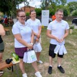 Trzy osoby w białych koszulkach stoją na trawie, trzymając torby. W tle drzewa i banner. Jasny, pogodny dzień.