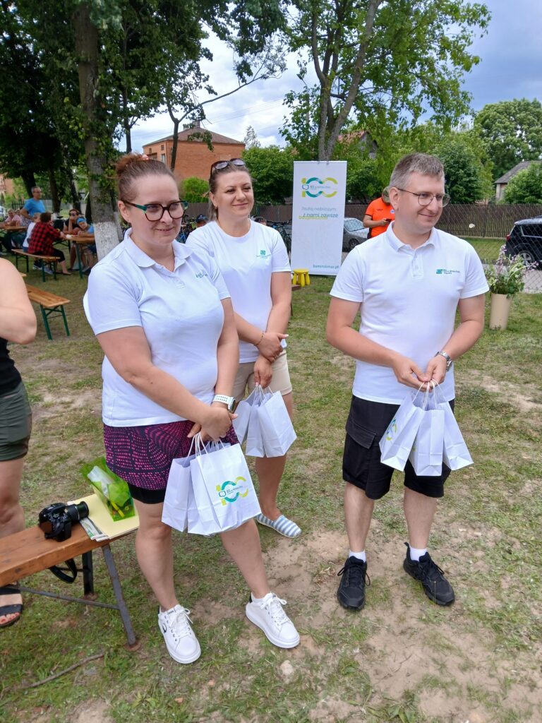 Trzy osoby w białych koszulkach stoją na trawie, trzymając torby. W tle drzewa i banner. Jasny, pogodny dzień.