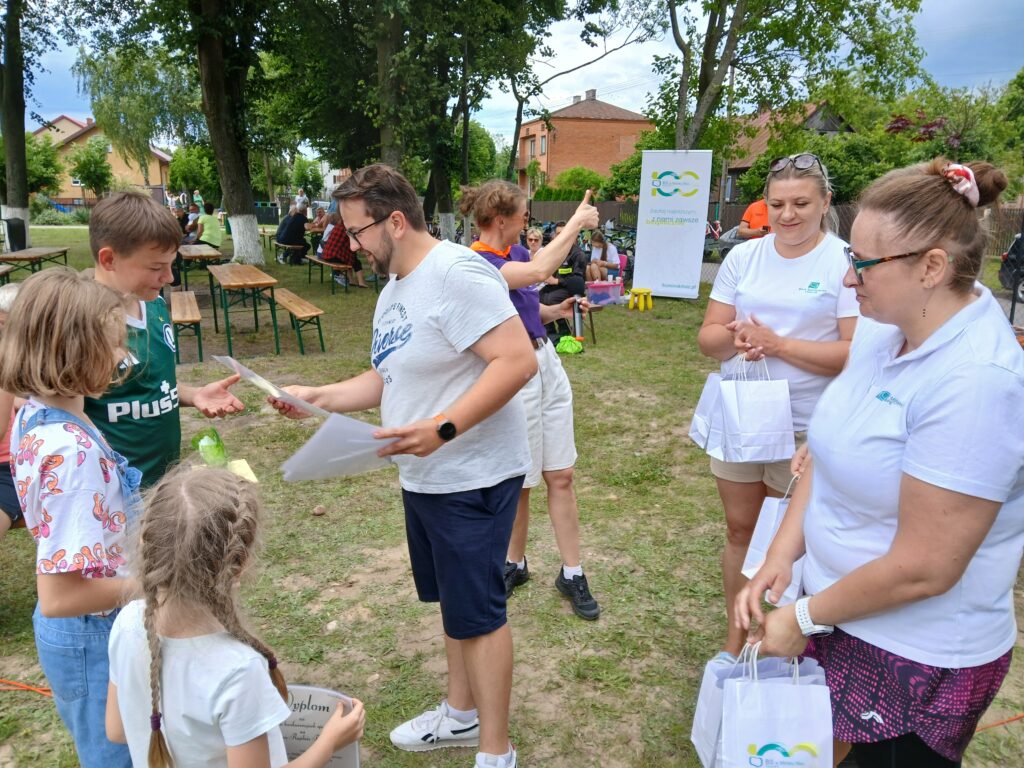 Na zdjęciu grupa osób na zewnątrz wymienia się dyplomami. Tło to drzewa i budynki. Ubrania są jasne, dominują białe i zielone kolory.
