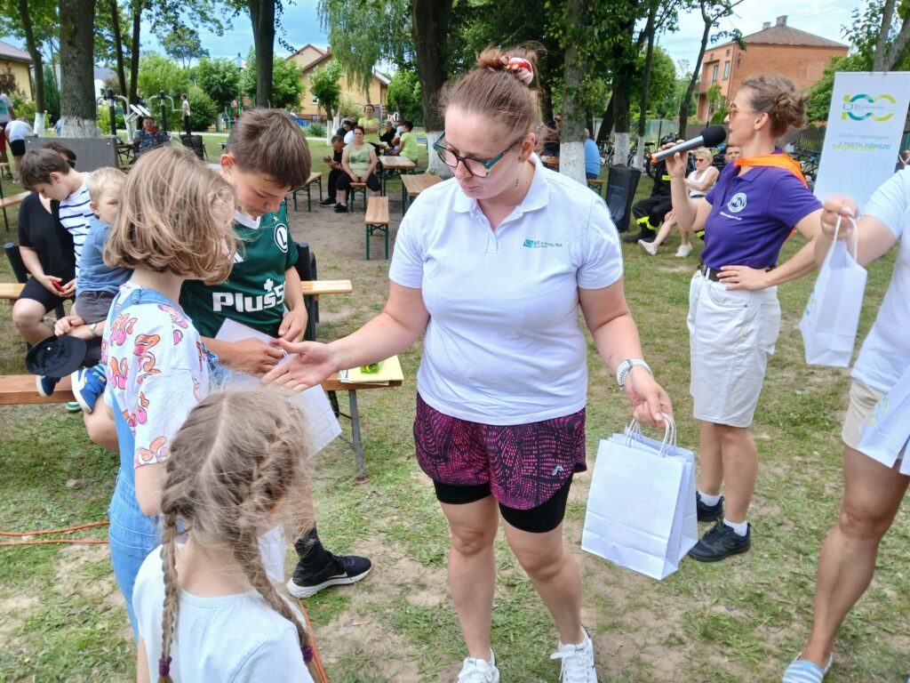 Na zdjęciu w parku kobieta w białej koszulce rozdaje dzieciom torby z prezentami. W tle ludzie siedzą na ławkach pod drzewami. Dzień jest pogodny z zielenią drzew i trawą.